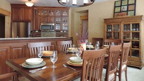A dining area with a wooden table set for six, surrounded by chairs. There is a chandelier above the table, and a cabinet with glass doors to the right.