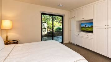 A hotel room with a bed, a television on a cabinet, a lamp, and a sliding glass door leading to a garden.