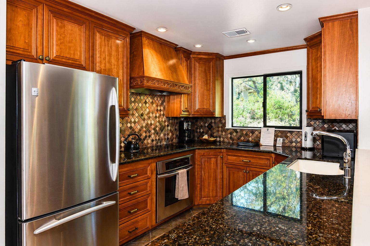 A kitchen with wooden cabinets, a stainless steel refrigerator, granite countertops, and a window with a view of greenery.