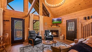 Interior acogedor de una cabaña de madera con chimenea, sofás de cuero, un televisor y un ventanal con vistas al exterior.