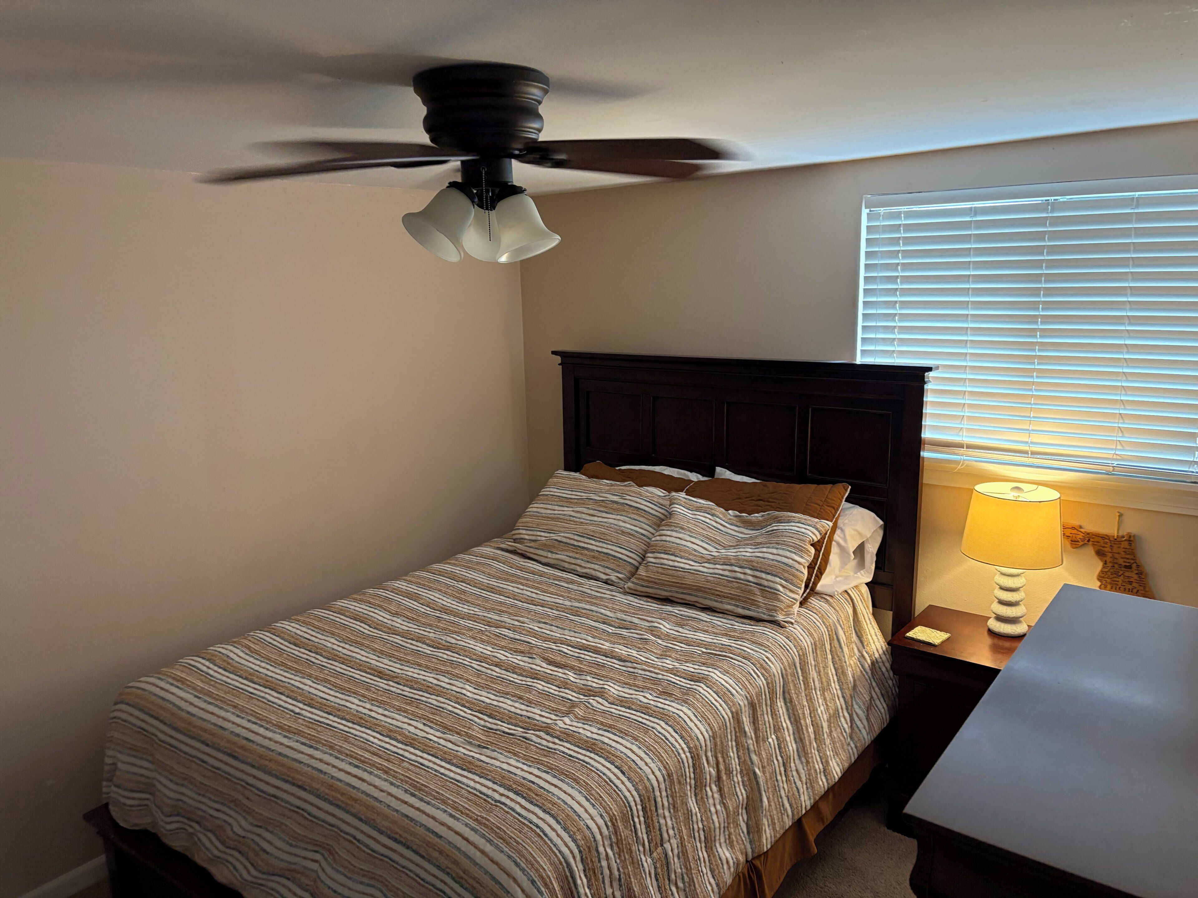 Een slaapkamer met een bed, een plafondventilator, een nachtkastje met lamp en een raam met jaloezieën.
