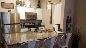 Una cocina con armarios blancos, un refrigerador de acero inoxidable, un microondas, una estufa y un lavamanos. Hay tres taburetes blancos y una planta en maceta.