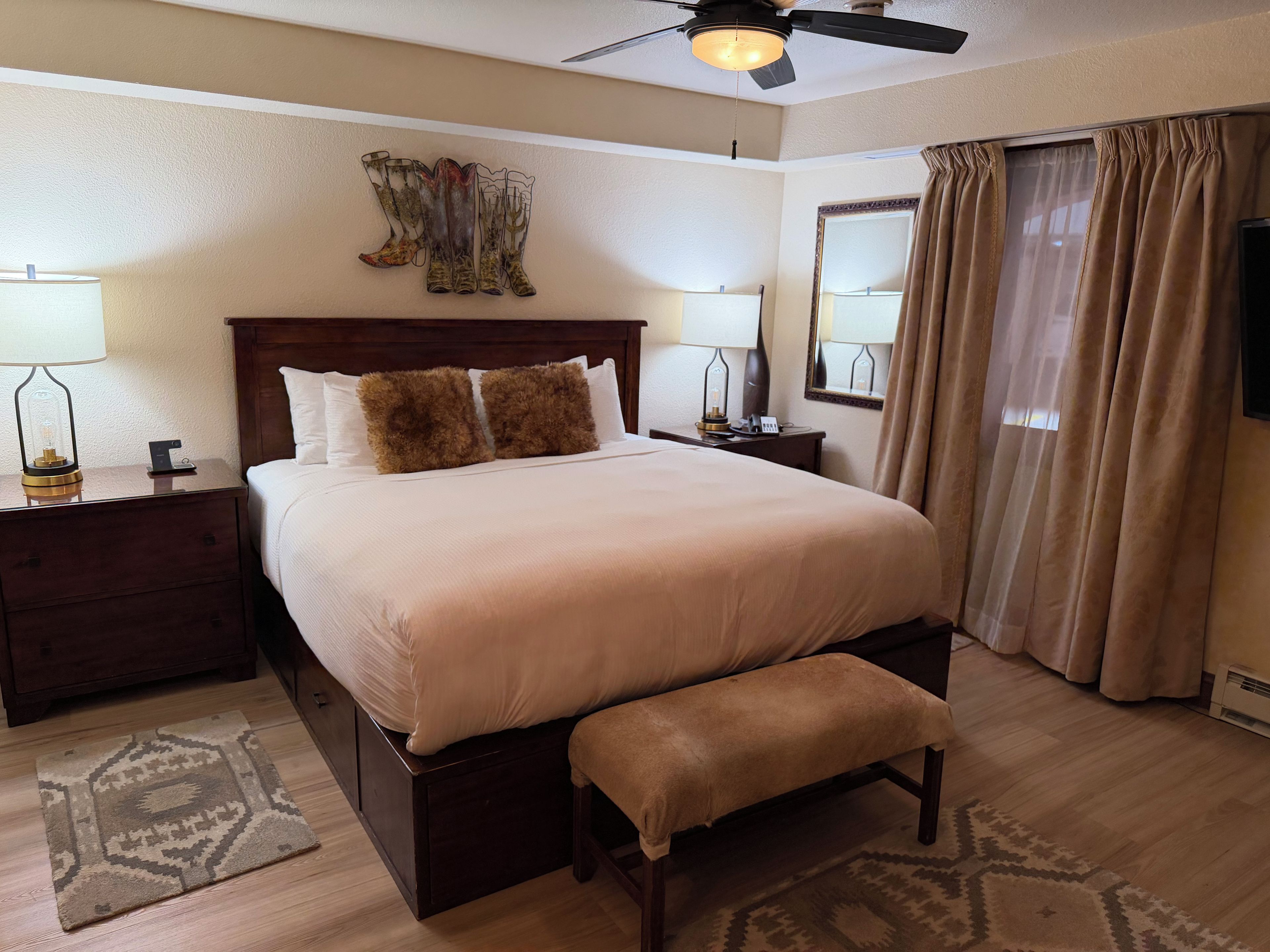 A neatly arranged bedroom with a large bed, two bedside tables with lamps, a ceiling fan, a large mirror, and a window with curtains.