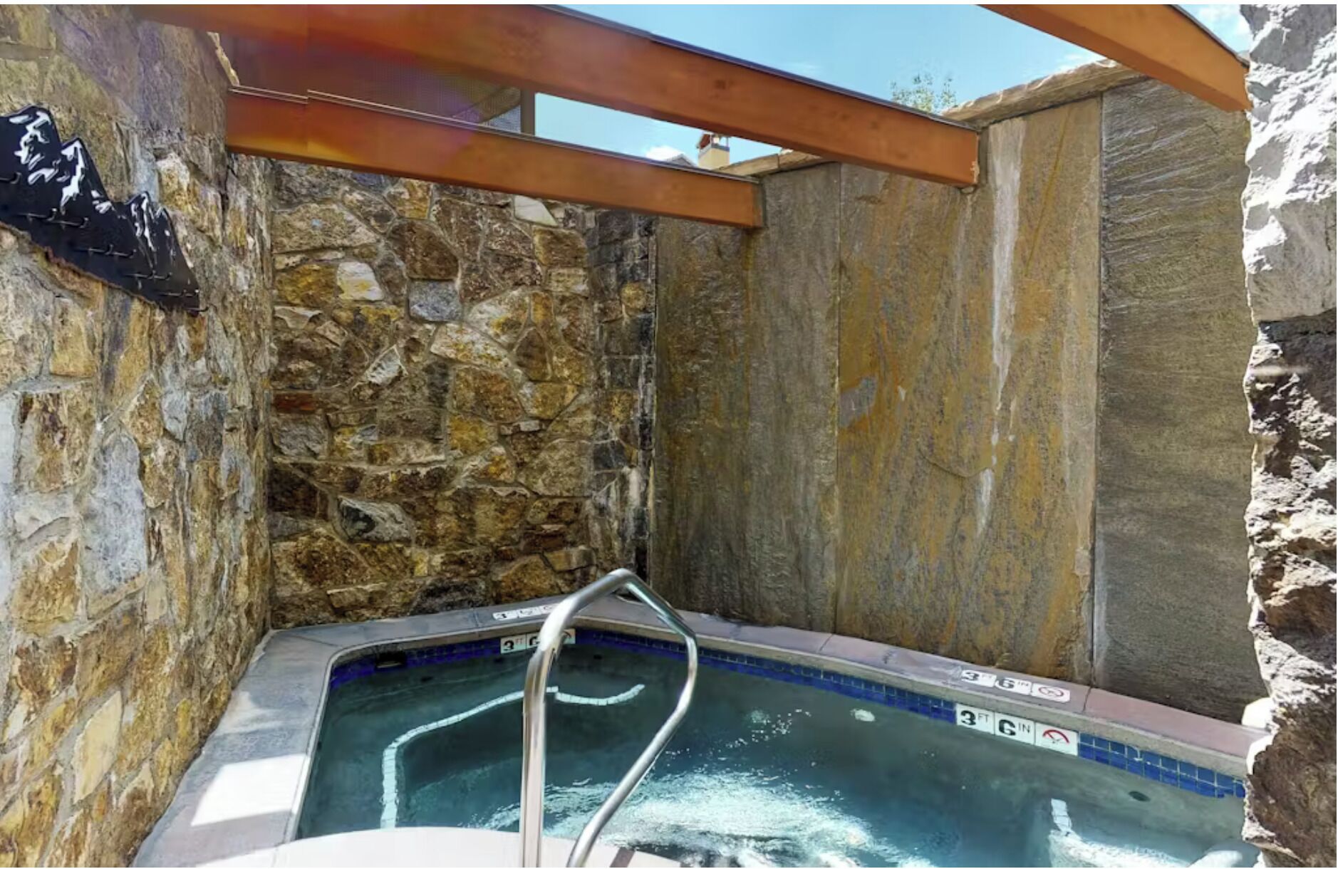 A stone-walled hot tub with a wooden roof structure, a metal ladder, and a tiled edge.