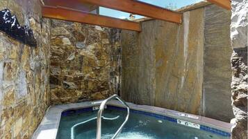 A stone-walled hot tub with a wooden roof structure, a metal ladder, and a tiled edge.