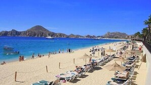 A beach with rows of sun loungers and umbrellas, a clear blue sea, and distant mountains. - UNBELIEVABLE VIEWS-EXTRA LARGE BALCONY FOUR LOUNGE CHAIRS 3 KING BEDS (Cabo San Lucas)