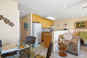 A compact kitchen with wooden cabinets, a refrigerator, microwave, and a dining area with a glass table and chairs.