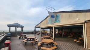 Une terrasse en bois avec des tables de pique-nique et des bancs, un espace de repos couvert, et un bâtiment au design ondulé portant l’enseigne « The Quarter Beach Restaurant and Bar ».