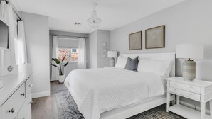 A modern bedroom with a large bed, bedside tables, a chandelier, and a view of the outside. - Newly renovated upscale Beach House  (Bay Head)