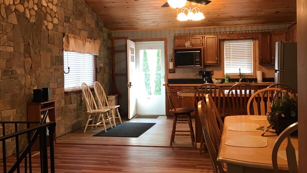 A spacious kitchen with wooden cabinets, a microwave, and a refrigerator. There is a dining area with a wooden table and chairs, and a stone wall with a window. - Adirondacks Family Retreat (Fort Ann)