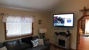 A living room with a leather sofa, a fireplace, a television, and a window with blinds. - Adirondacks Family Retreat (Fort Ann)