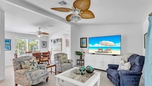 Ein großzügiges Wohnzimmer mit einem Fernseher im Strandhaus-Stil, zwei Sesseln, einem Couchtisch und Deckenventilatoren.