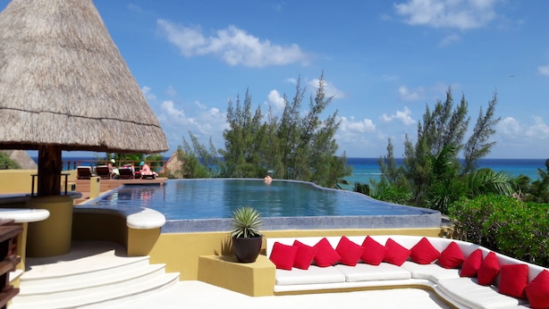 A pool area with a thatched roof structure, a white sofa with red cushions, and a view of the ocean.