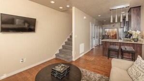 A modern living room with a flat-screen TV, a coffee table with books, a staircase leading to a kitchen area with dark wood cabinets and a marble countertop. - Springdale Townhome 1 (Springdale)