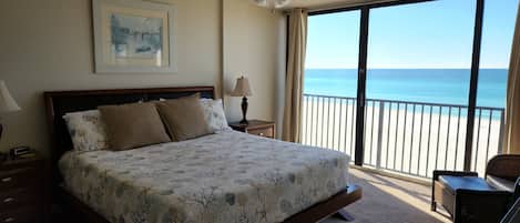 A bedroom with a bed, a nightstand, a lamp, a ceiling fan, a window with a view of the beach, and a chair.