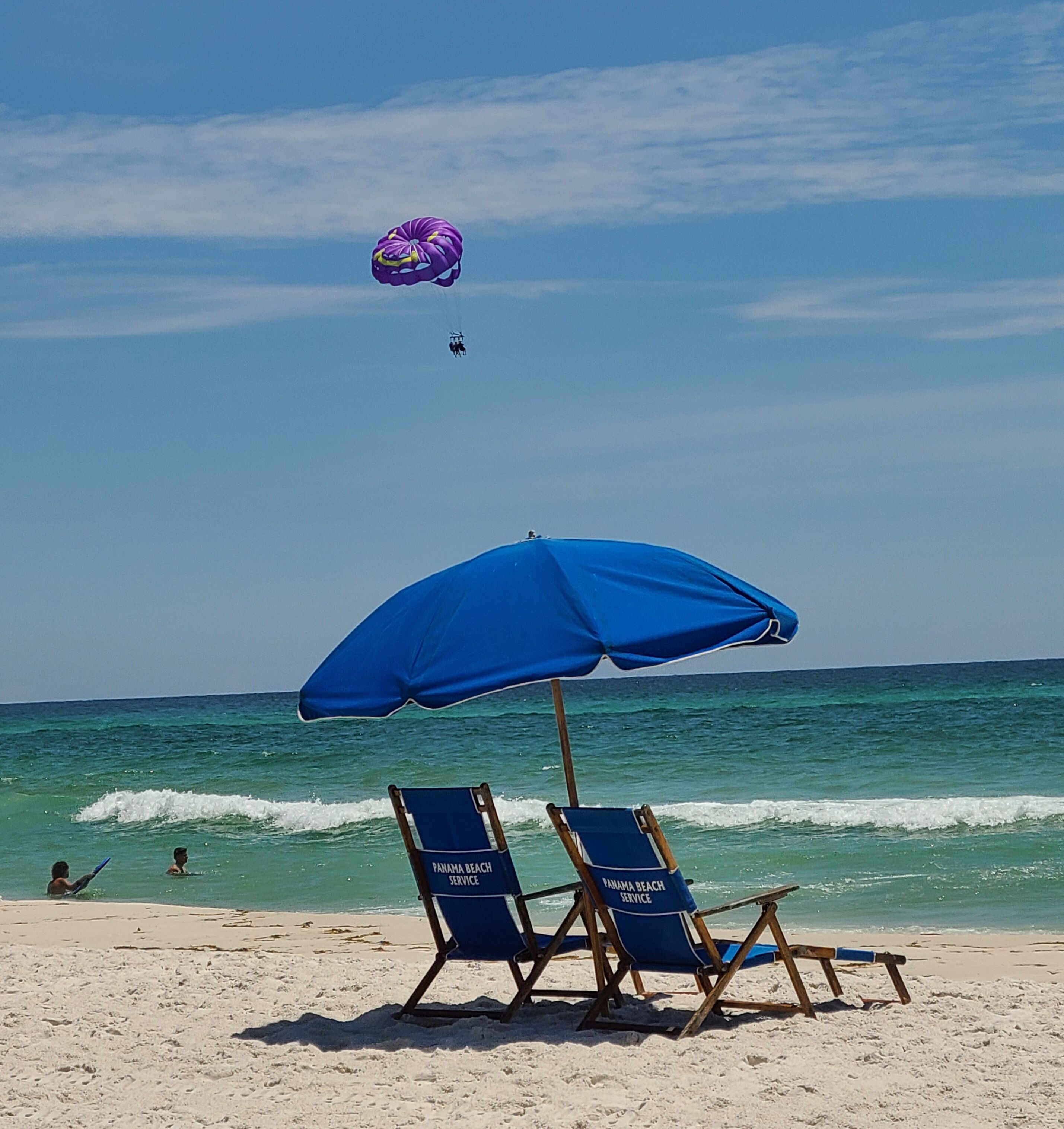 Una spiaggia con due sdraio blu e un ombrellone blu, un paracadute viola sullo sfondo e l'oceano.
