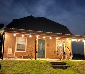Una cabaña de madera con dos ruedas grandes al frente, una puerta verde y un pequeño porche con una silla y una mesa.
