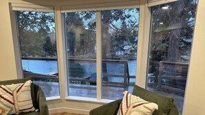 Una habitación con dos sillones verdes, cada uno con un cojín estampado, frente a ventanas amplias que dan a una terraza de madera y una masa de agua.