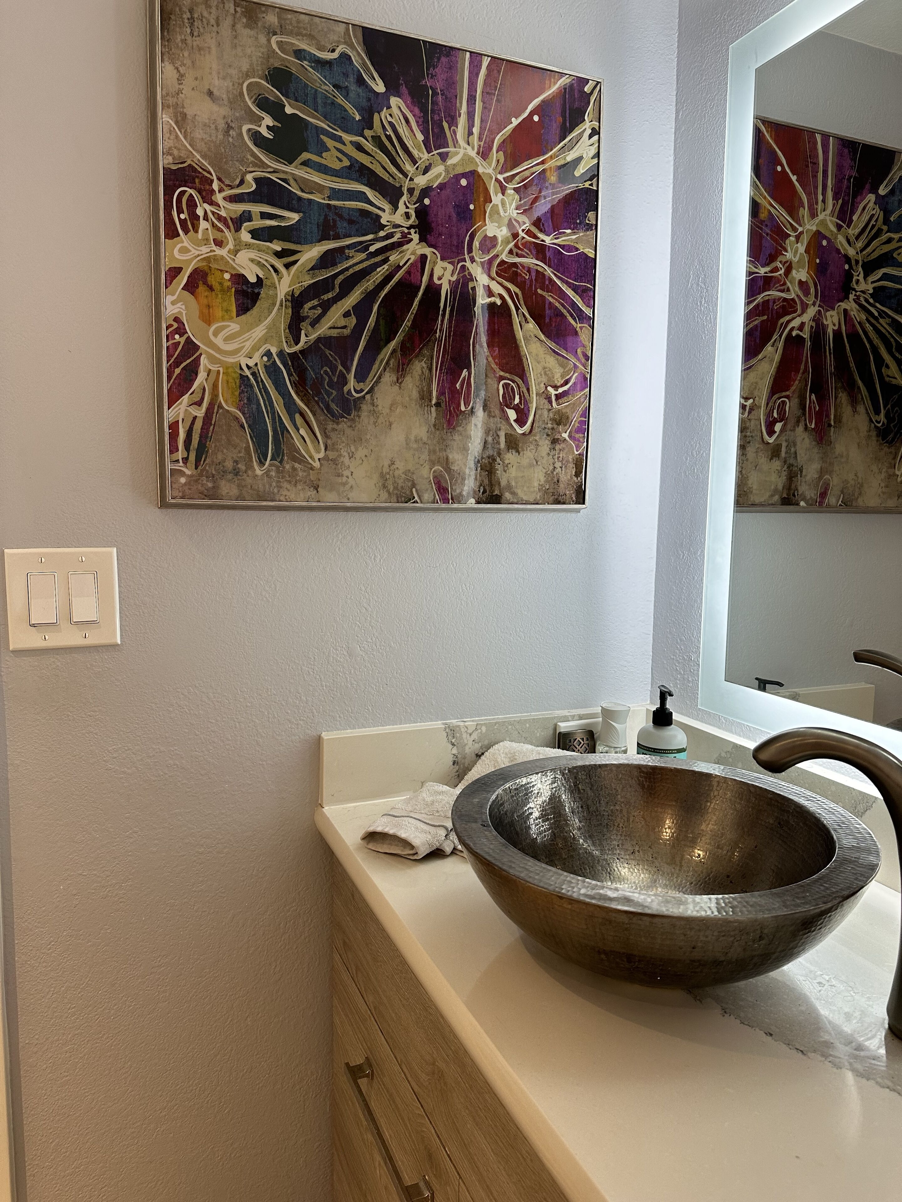 Un baño con un cuadro abstracto grande, un tocador de madera y un lavabo sobre encimera.