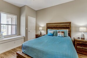 Une chambre avec un grand lit, une fenêtre avec des stores, une tête de lit en bois, deux tables de chevet avec des lampes et une petite table de nuit avec une bouilloire.