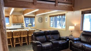 A cozy living room with a leather sofa and wooden chairs, a kitchen in the background, and large windows. - Deer Creek Cabin Hot Tub, BBQ Grill, Fire Pit, 20 min Yellowstone (Island Park)
