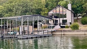 Uma propriedade à beira de um lago, com um píer coberto, uma casa moderna com grandes janelas e um morro arborizado ao fundo.