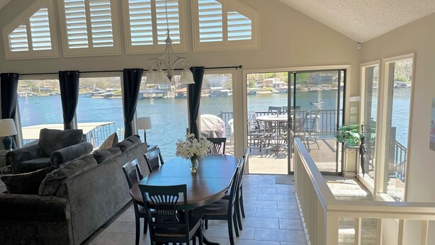 Uma sala de jantar com mesa e cadeiras, um sofá e vista para um lago através de grandes janelas.