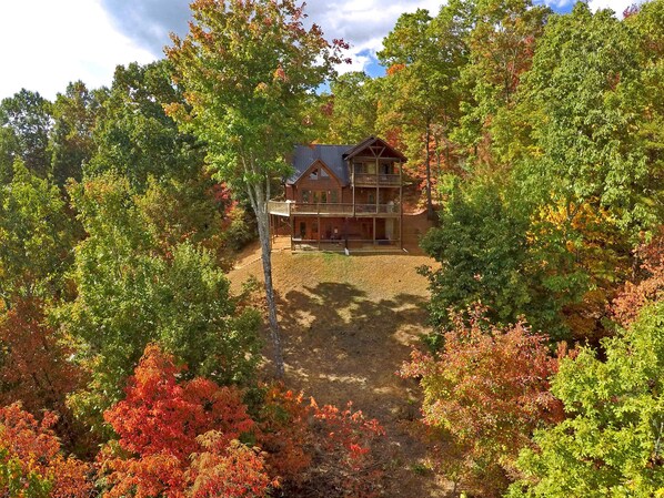 A two-story log cabin with a balcony, surrounded by dense forest with autumn foliage.