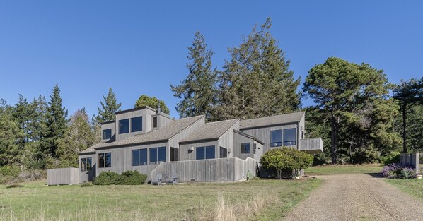 A modern two-story house with a sloped roof, large windows, and a wooden fence. - Abalone Point:  Oceanside Panoramic View Home (Sea Ranch)