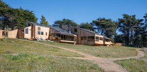 A modern wooden cabin with multiple levels and large windows, situated on a grassy hillside with a clear sky. - Abalone Point:  Oceanside Panoramic View Home (Sea Ranch)