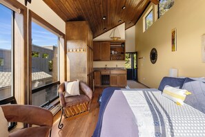A bedroom with a large bed, a sofa, and a chair. There is a kitchen area with wooden cabinets and a window overlooking a garden. - Abalone Point:  Oceanside Panoramic View Home (Sea Ranch)