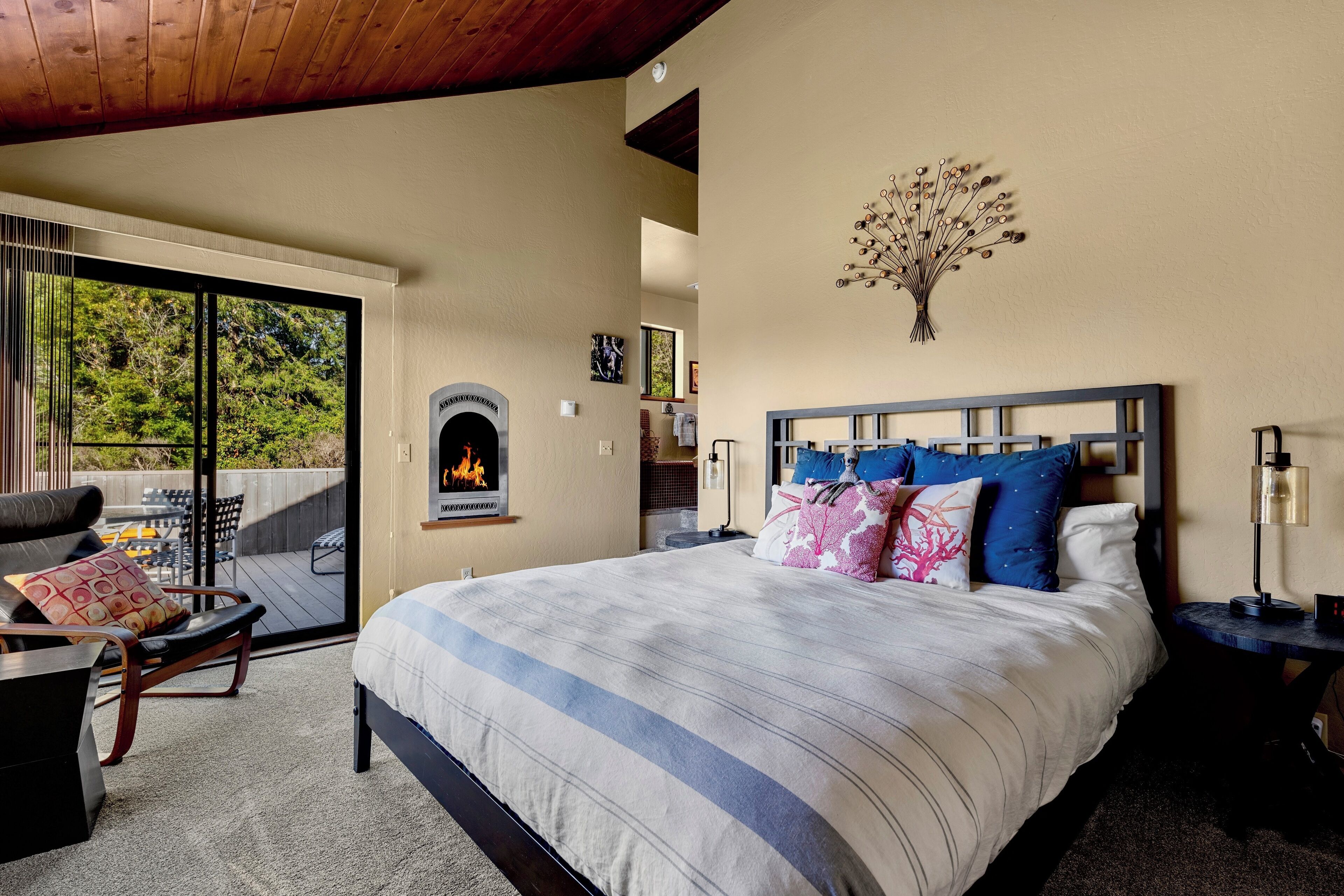 A bedroom with a large bed, a fireplace, a balcony with outdoor furniture, and a tree wall art piece.