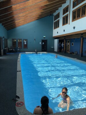 An indoor swimming pool with clear blue water, wooden ceiling beams, and a blue wall with windows.