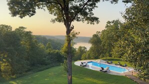 Una piscina con lettini, circondata da vegetazione rigogliosa e vista su un lago.