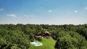 Una casa grande rodeada de un bosque frondoso, con piscina y mobiliario exterior.