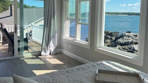 Een slaapkamer met een groot bed, twee ramen met uitzicht op zee, een plafondventilator en een schuifdeur naar een balkon.