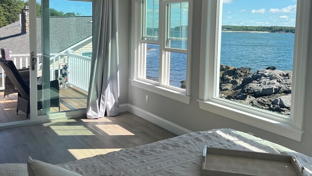 A bedroom with a large bed, two windows with ocean views, a ceiling fan, and a sliding glass door leading to a balcony.