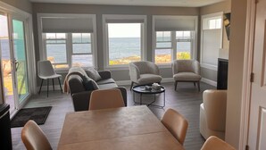 A spacious room with a large table and several chairs, large windows with ocean views, and a door leading to another area.