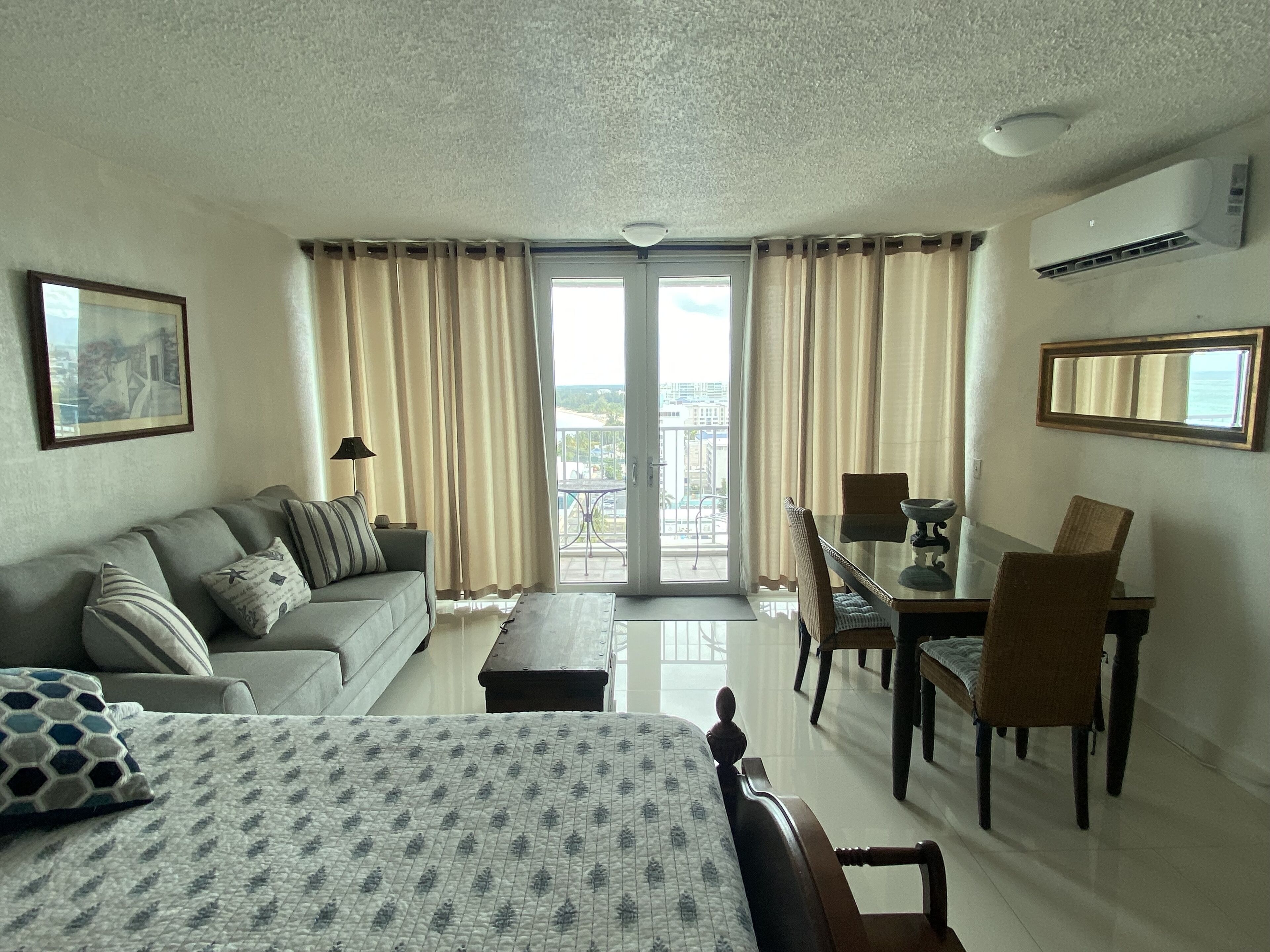 A hotel room with a bed, a sofa, a dining table with chairs, a balcony with curtains, and a framed picture on the wall.