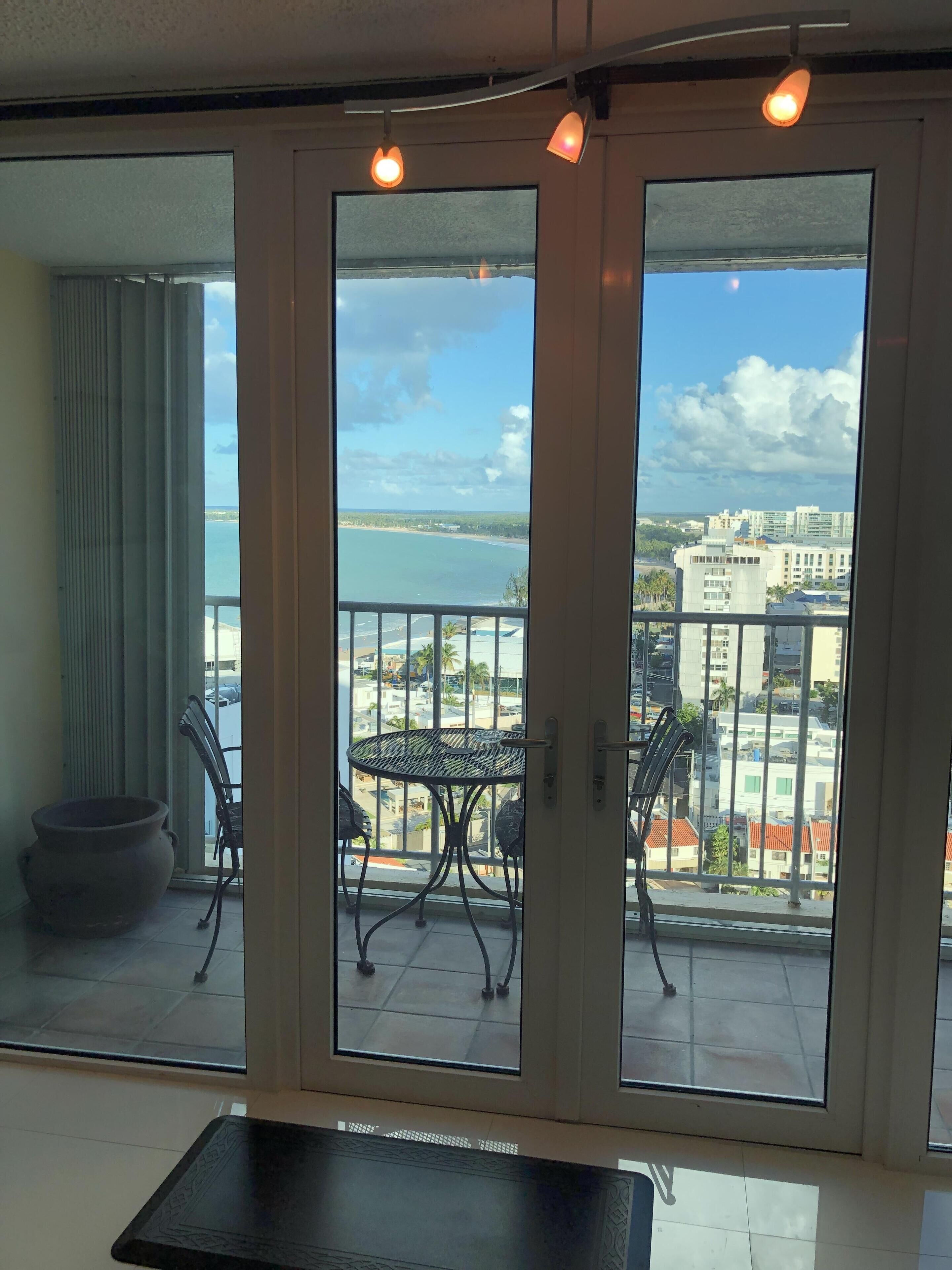 A balcony with a table and chairs, a view of the sea and buildings, and a pot on the floor.
