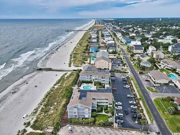 An aerial view of a coastal town with a beach, a pier, and a row of multi-story buildings with parking and a swimming pool.