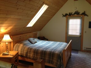 Une chambre avec un plafond en pente, un lit recouvert d’une couette à carreaux, une lampe de chevet et une fenêtre avec des stores.