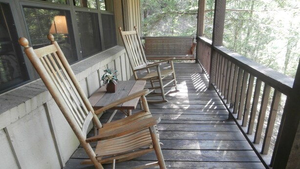 A wooden porch with two rocking chairs and a small table, overlooking a forested area.