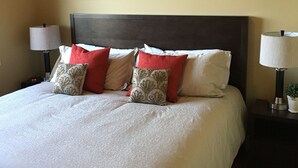 A neatly made bed with white bedding and decorative pillows, flanked by two bedside tables with lamps and a small plant. - "Quails Run Retreat" on Bear Mountain  (Victoria)