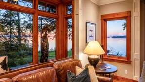 A cozy room with a leather sofa, a lamp, and large windows offering a view of a lake and trees. - Orcas Waterfront Custom Home, 180 Degree Summer Sunset & Sunrise,  Sleeps 12 (Eastsound)