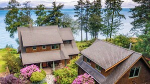 A two-story house with a stone chimney, a garage, and a well-maintained garden with pink flowers. - Orcas Waterfront Custom Home, 180 Degree Summer Sunset & Sunrise,  Sleeps 12 (Eastsound)