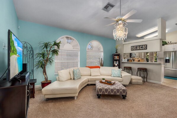 A living room with a sectional sofa, a TV, a ceiling fan, and a kitchen area with a sign that reads 'BEACH'. - Heated Pool! 5 Minute Bike To Beach, Dining, Mercato! Bikes Included (Naples)