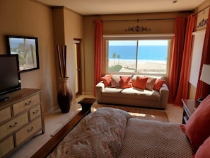 A bedroom with a bed, a sofa, a TV, a dresser, and a window with a beach view.