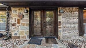 A stone and brick exterior with a dark wooden door, two lantern-style lights, and a metal wind chime. - Resort Quality 4B Home With Private Pool In Beautiful Hill Country (New Braunfels)