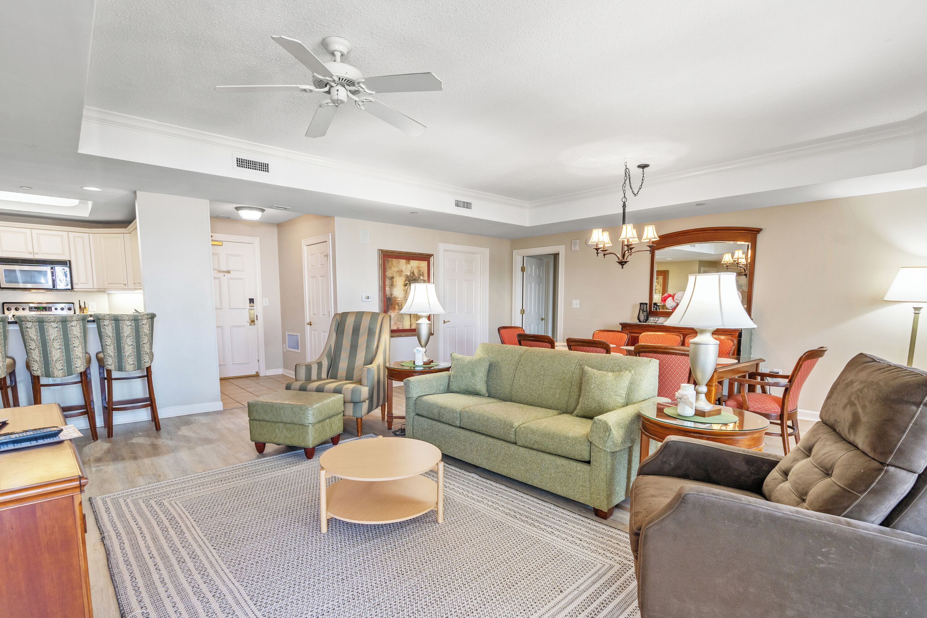 A spacious living room with a green sofa, a brown armchair, and a round coffee table. There is a kitchen area with a microwave and chairs.